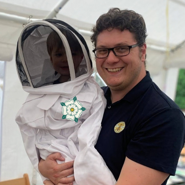 Richard Senior smiling and holding a young visitor dressed in a white beekeeping suit and veil